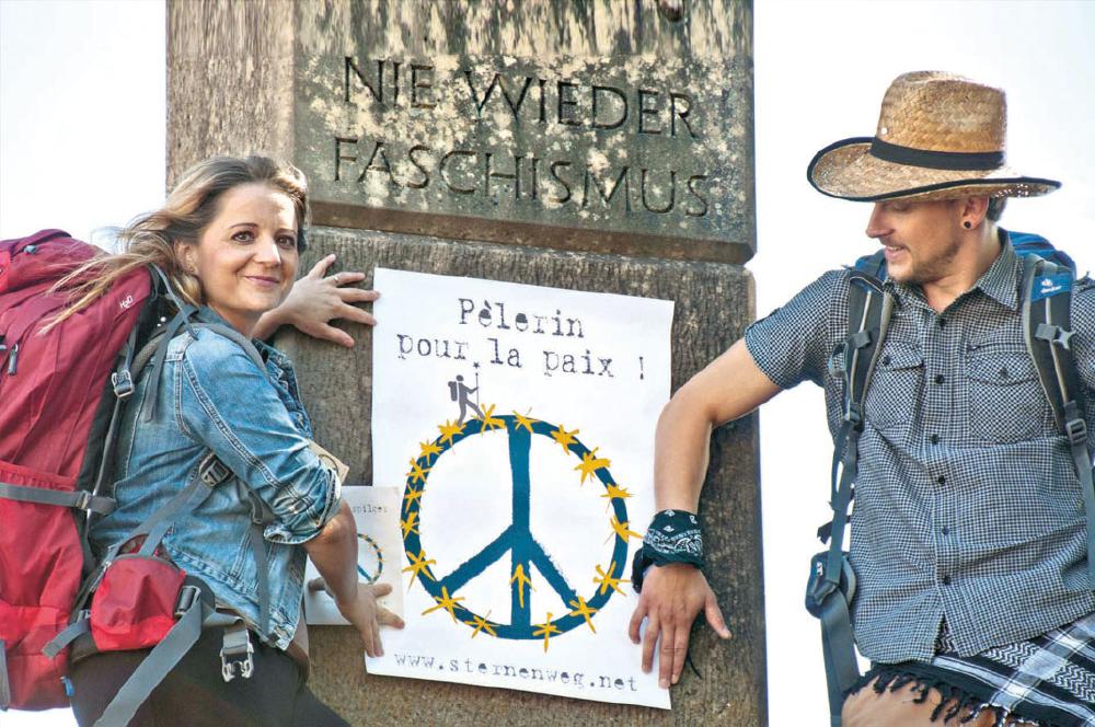 Zwei Pilger vor Kriegsmahnmal zeigen ein Plakat