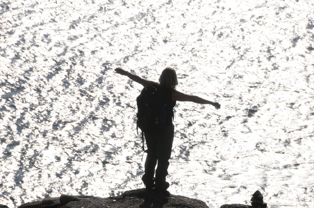 Frau mit ausgebreiteten Armen auf Felsvorsprung am windbewegten Wasser