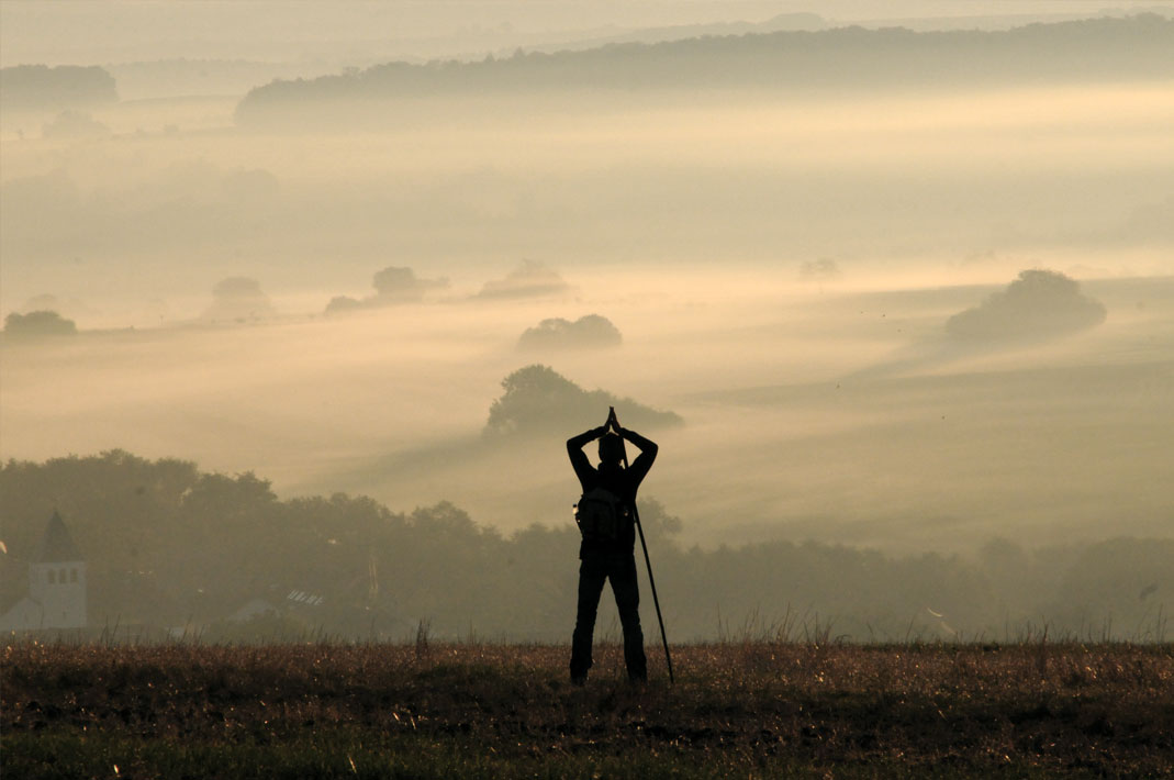 Mann mit Wanderstock und gefalteten Händen über dem Kopf schaut auf nebelschwangere Landschaft